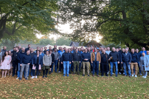 Große Gruppe von etwa 60 Personen unterschiedlichen Alters steht auf einer Wiese unter Bäumen mit herbstlichem Laub. Die meisten tragen lässige Freizeitkleidung, einige wenige traditionelle Trachten. Im Hintergrund sind Häuser und Bäume zu sehen. Das Bild vermittelt ein Gemeinschaftsgefühl und passt zum Team der dierckgroup.