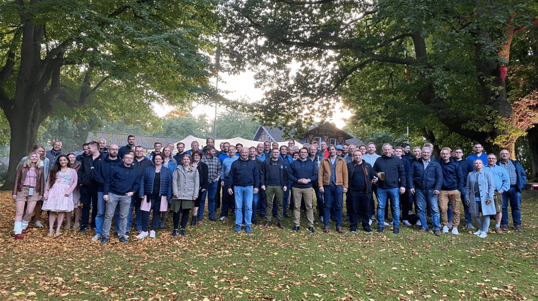 Große Gruppe von etwa 60 Personen unterschiedlichen Alters steht auf einer Wiese unter Bäumen mit herbstlichem Laub. Die meisten tragen lässige Freizeitkleidung, einige wenige traditionelle Trachten. Im Hintergrund sind Häuser und Bäume zu sehen. Das Bild vermittelt ein Gemeinschaftsgefühl und passt zum Team der dierckgroup.