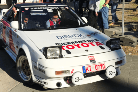 Weißer Toyota-Rennwagen mit Rallye-Aufklebern, darunter „dierckgroup“, „Mickhams Oils“ und „Motorsportfreunde Kiel“, zwei Personen im Fahrzeug, umgeben von Zuschauern auf gepflastertem Platz bei sonnigem Wetter.