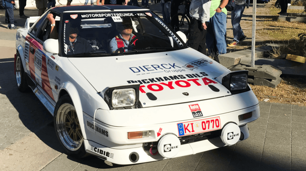 Weißer Toyota-Rennwagen mit Rallye-Aufklebern, darunter „dierckgroup“, „Mickhams Oils“ und „Motorsportfreunde Kiel“, zwei Personen im Fahrzeug, umgeben von Zuschauern auf gepflastertem Platz bei sonnigem Wetter.