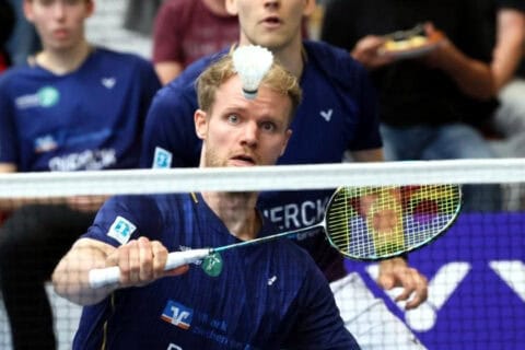 Zwei Männer in blauen Sportshirts spielen Badminton in einer Halle, einer konzentriert beim Schlag auf den Federball, der knapp über dem Netz schwebt, im Hintergrund Zuschauer auf der Tribüne.