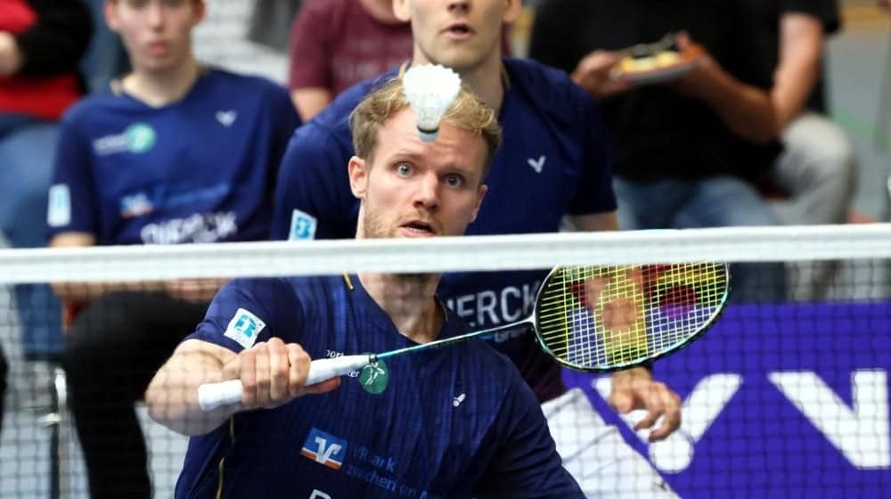 Zwei Männer in blauen Sportshirts spielen Badminton in einer Halle, einer konzentriert beim Schlag auf den Federball, der knapp über dem Netz schwebt, im Hintergrund Zuschauer auf der Tribüne.