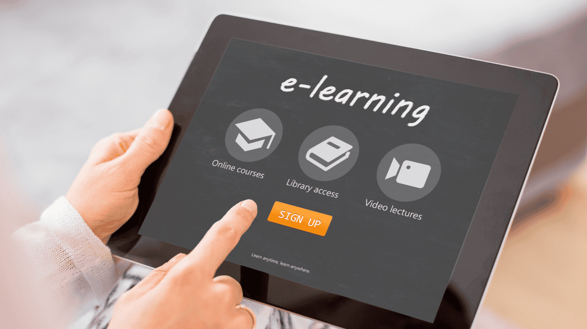 Tablet mit e-learning Startseite, darauf Symbole für Online-Kurse, Bibliothekszugang und Video-Vorlesungen, darunter ein orangefarbener Button mit der Aufschrift „Sign Up“, gehalten von einer Person mit hellem Pullover, die mit dem Finger auf den Bildschirm zeigt.