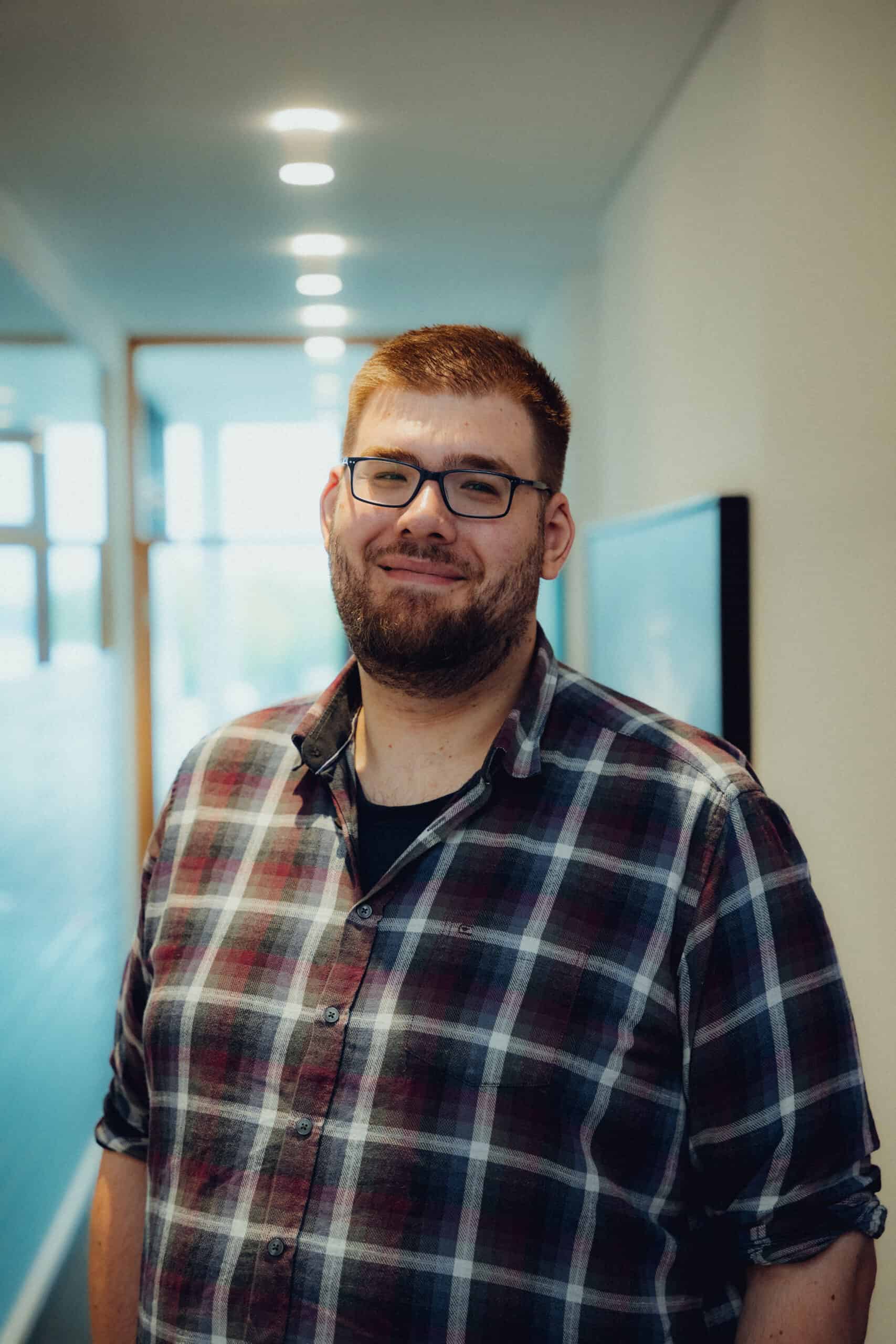 Junger Mann mit Brille und kurzem Bart trägt kariertes Hemd mit hochgekrempelten Ärmeln, steht lächelnd in einem hellen Flur mit Deckenbeleuchtung, unscharfer Hintergrund mit Fenstern und Türrahmen.
