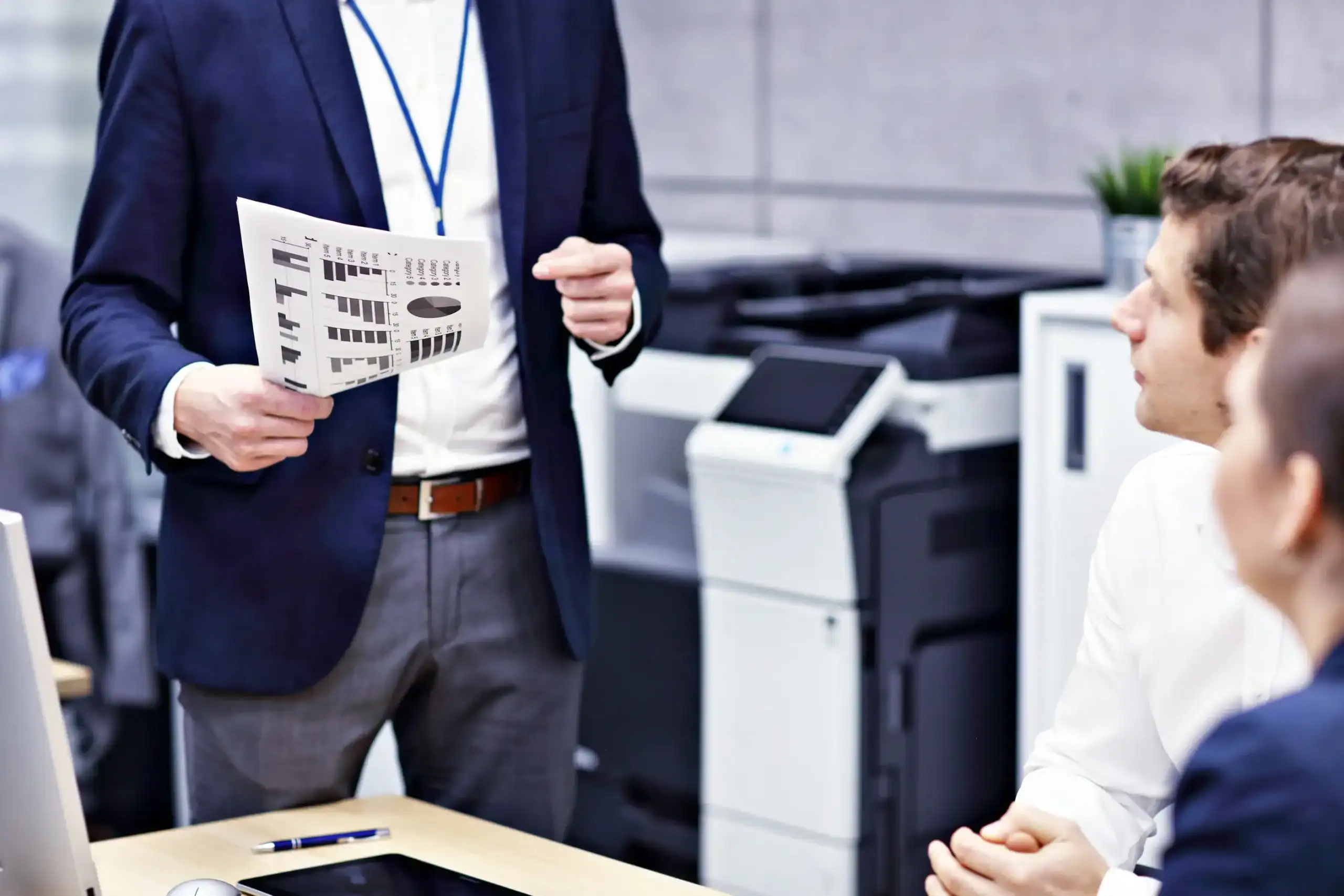 Mann in Business-Kleidung hält ein Blatt mit Diagrammen und präsentiert vor zwei sitzenden Kollegen in einem modernen Büro mit Drucker im Hintergrund, symbolisiert IT-Beratung und Kommunikationstechnik bei der dierckgroup.