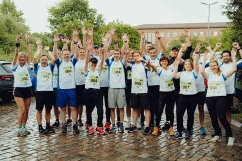 Gruppe von 17 Läufern in weißen und blauen Sportshirts mit Startnummern, die auf nassem Kopfsteinpflaster stehen und die Arme jubelnd in die Luft strecken, im Hintergrund Bäume und ein Backsteingebäude, symbolisiert Teamgeist und Zusammenhalt bei einem Firmenlauf der dierckgroup.