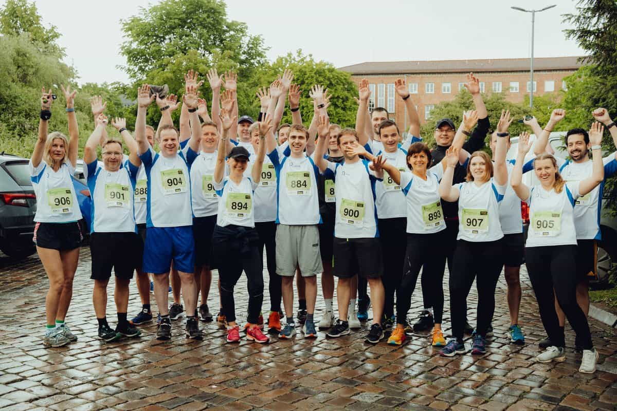 Gruppe von 17 Läufern in weißen und blauen Sportshirts mit Startnummern, die auf nassem Kopfsteinpflaster stehen und die Arme jubelnd in die Luft strecken, im Hintergrund Bäume und ein Backsteingebäude, symbolisiert Teamgeist und Zusammenhalt bei einem Firmenlauf der dierckgroup.