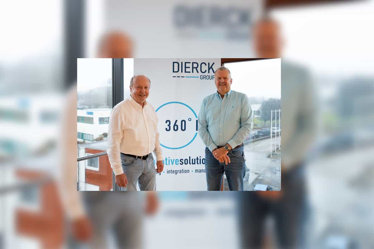 Zwei Männer stehen vor einem Fenster mit Blick auf ein Gewerbegebiet. Im Hintergrund ein Banner mit dem Schriftzug „dierckgroup“ und „360° innovative solutions“. Die Männer tragen Hemden, einer weiß, der andere hellblau, und lächeln in die Kamera.
