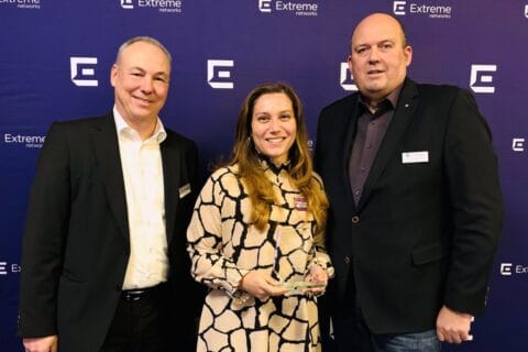Drei Personen vor blauem Hintergrund mit mehrfach wiederholtem Logo von Extreme Networks. Die Frau in der Mitte hält einen gläsernen Award in den Händen und trägt ein helles, gemustertes Kleid. Die beiden Männer links und rechts tragen dunkle Anzüge und Hemden. Die Szene wirkt wie eine Auszeichnung oder Preisverleihung im IT-Umfeld.