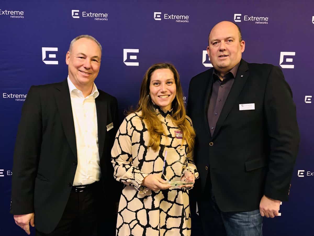 Drei Personen vor blauem Hintergrund mit mehrfach wiederholtem Logo von Extreme Networks. Die Frau in der Mitte hält einen gläsernen Award in den Händen und trägt ein helles, gemustertes Kleid. Die beiden Männer links und rechts tragen dunkle Anzüge und Hemden. Die Szene wirkt wie eine Auszeichnung oder Preisverleihung im IT-Umfeld.