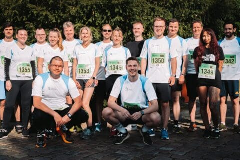 Gruppenfoto von 15 Personen in Sportkleidung mit Laufnummern, die vor einem grünen Hintergrund im Freien stehen. Die meisten tragen weiße Shirts mit blauen Ärmeln, einige haben schwarze oder dunkle Sportkleidung. Die Szene vermittelt Teamgeist und Aktivität, passend zu den Themen Digitalisierung und Workplace Solutions der dierckgroup.