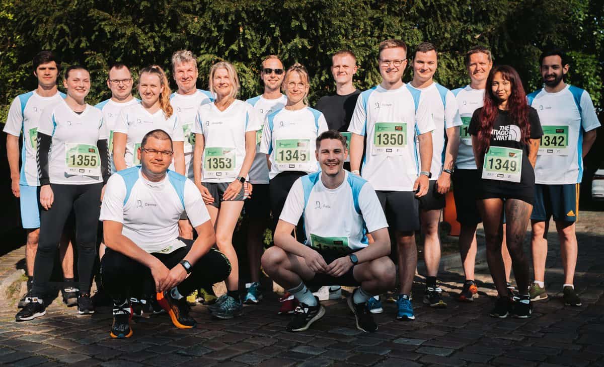 Gruppenfoto von 15 Personen in Sportkleidung mit Laufnummern, die vor einem grünen Hintergrund im Freien stehen. Die meisten tragen weiße Shirts mit blauen Ärmeln, einige haben schwarze oder dunkle Sportkleidung. Die Szene vermittelt Teamgeist und Aktivität, passend zu den Themen Digitalisierung und Workplace Solutions der dierckgroup.
