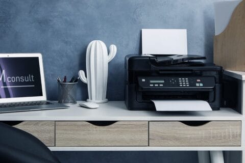 Moderner Arbeitsplatz mit grauem Schreibtisch vor blauer Wand, darauf ein Laptop mit geöffneter Webseite "HM consult", eine weiße Kaktus-Dekoration, ein Stiftehalter mit Stiften, eine weiße Computermaus und ein Multifunktionsdrucker mit Papier im Einzug. Rechts auf dem Schreibtisch ein hölzernes Ablagesystem mit Ordnern.