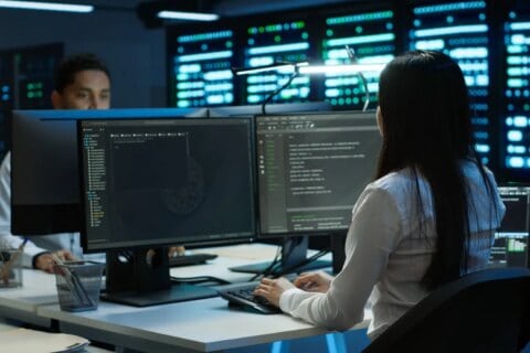 Junge IT-Programmiererin sitzt an einem Schreibtisch mit zwei großen Monitoren, auf denen Programmiercode und Dateistrukturen zu sehen sind, im Hintergrund Serverracks mit leuchtenden Kontrollleuchten, weitere Person am Arbeitsplatz erkennbar, moderne IT-Umgebung passend zu IT Solutions, Managed Services und Datacenter Solutions.