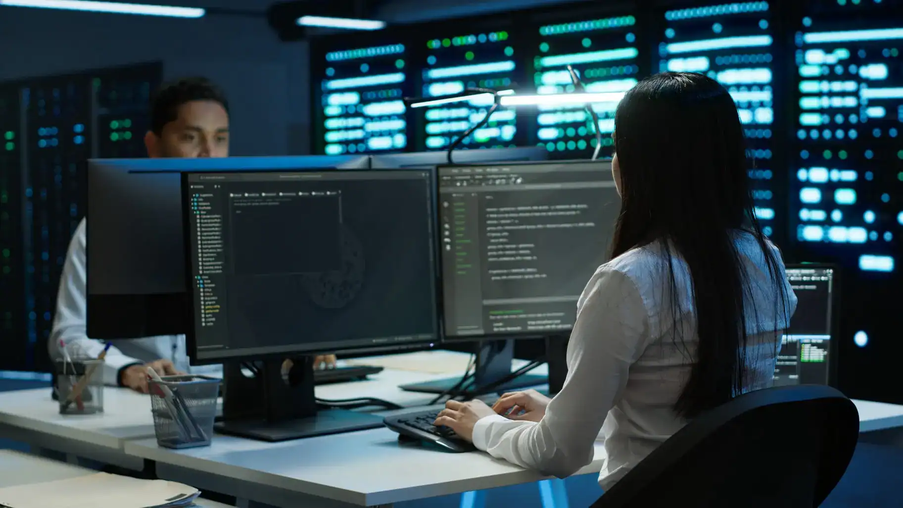 Junge IT-Programmiererin sitzt an einem Schreibtisch mit zwei großen Monitoren, auf denen Programmiercode und Dateistrukturen zu sehen sind, im Hintergrund Serverracks mit leuchtenden Kontrollleuchten, weitere Person am Arbeitsplatz erkennbar, moderne IT-Umgebung passend zu IT Solutions, Managed Services und Datacenter Solutions.
