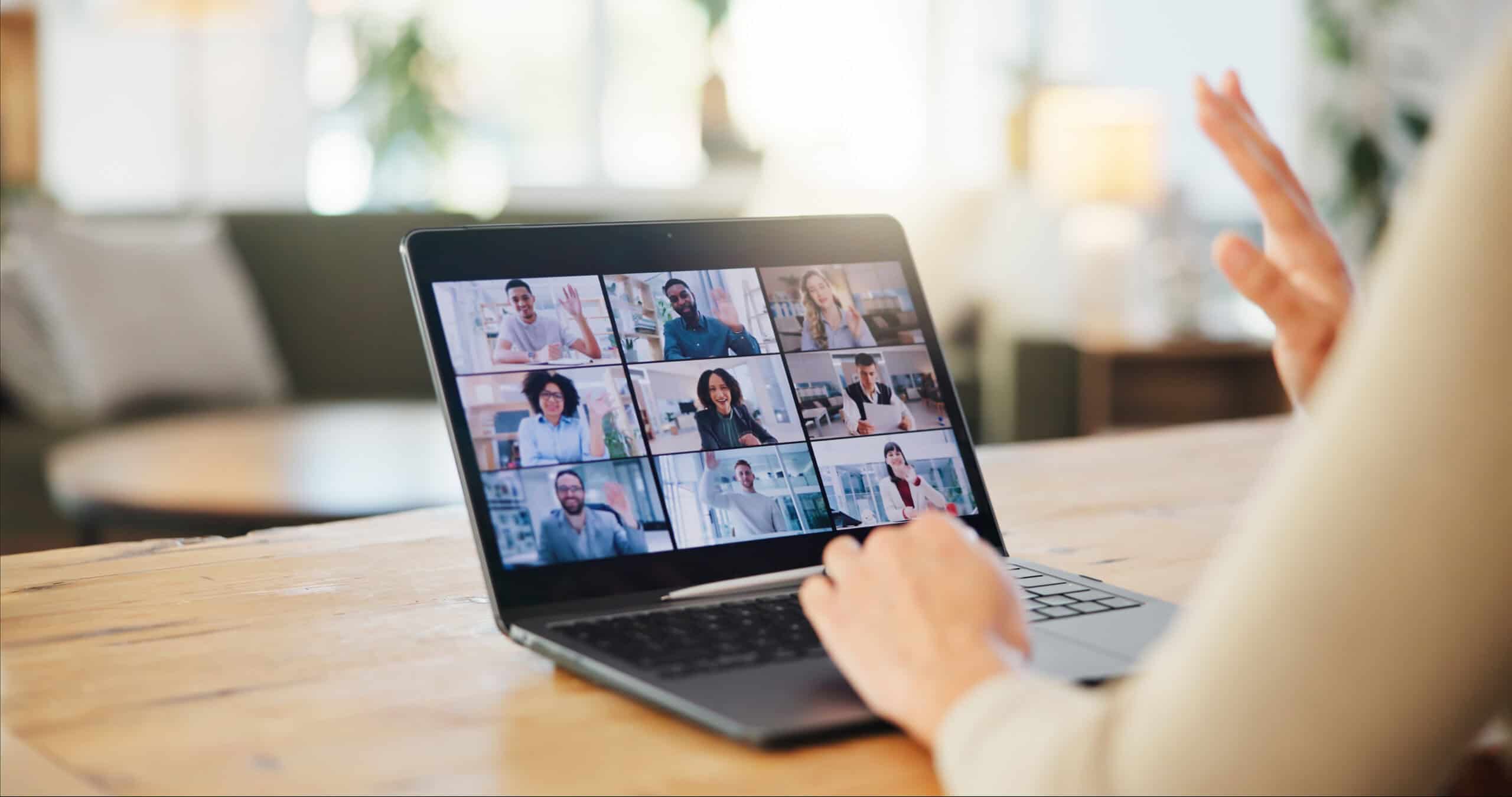 Laptop auf Holztisch mit Videokonferenz, neun Teilnehmer in einzelnen Fenstern, Personen winken und begrüßen sich, helle, freundliche Arbeitsumgebung, symbolisiert digitale Kommunikation und Zusammenarbeit.