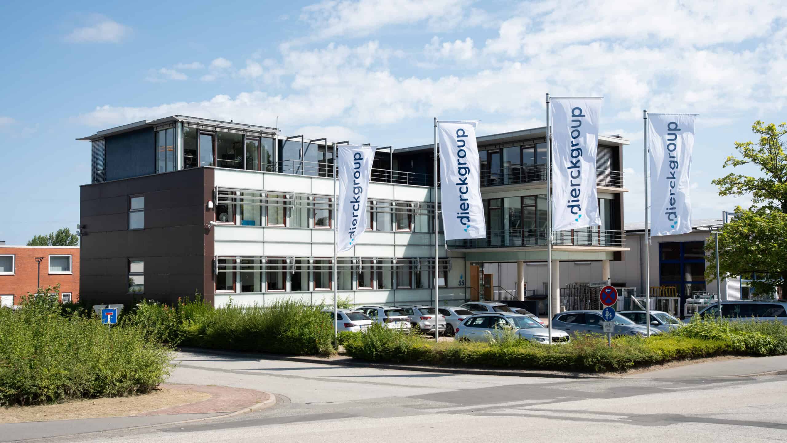 Modernes Bürogebäude der dierckgroup mit drei weißen Fahnen, auf denen der Firmenname steht, umgeben von Parkplätzen und Grünflächen, unter blauem Himmel.