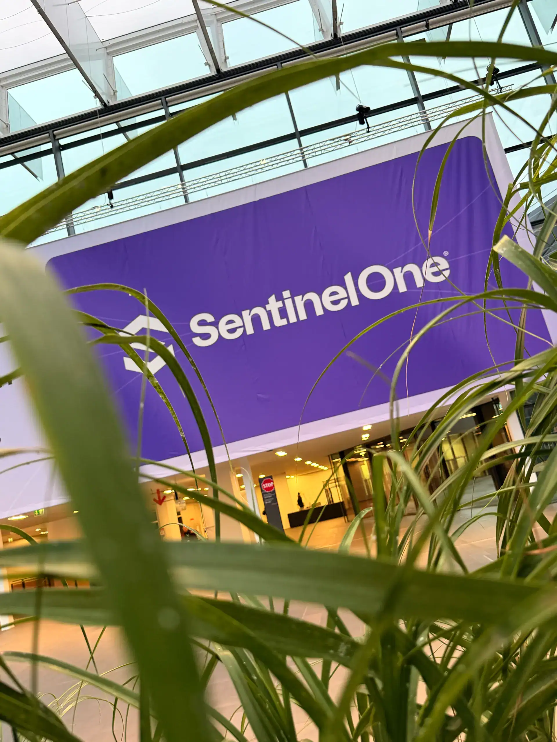 Großes lila Banner mit weißer Aufschrift „SentinelOne“ in moderner Halle mit Glasdach, im Vordergrund grüne Pflanzen, im Hintergrund Empfangsbereich mit Beleuchtung und Personen.