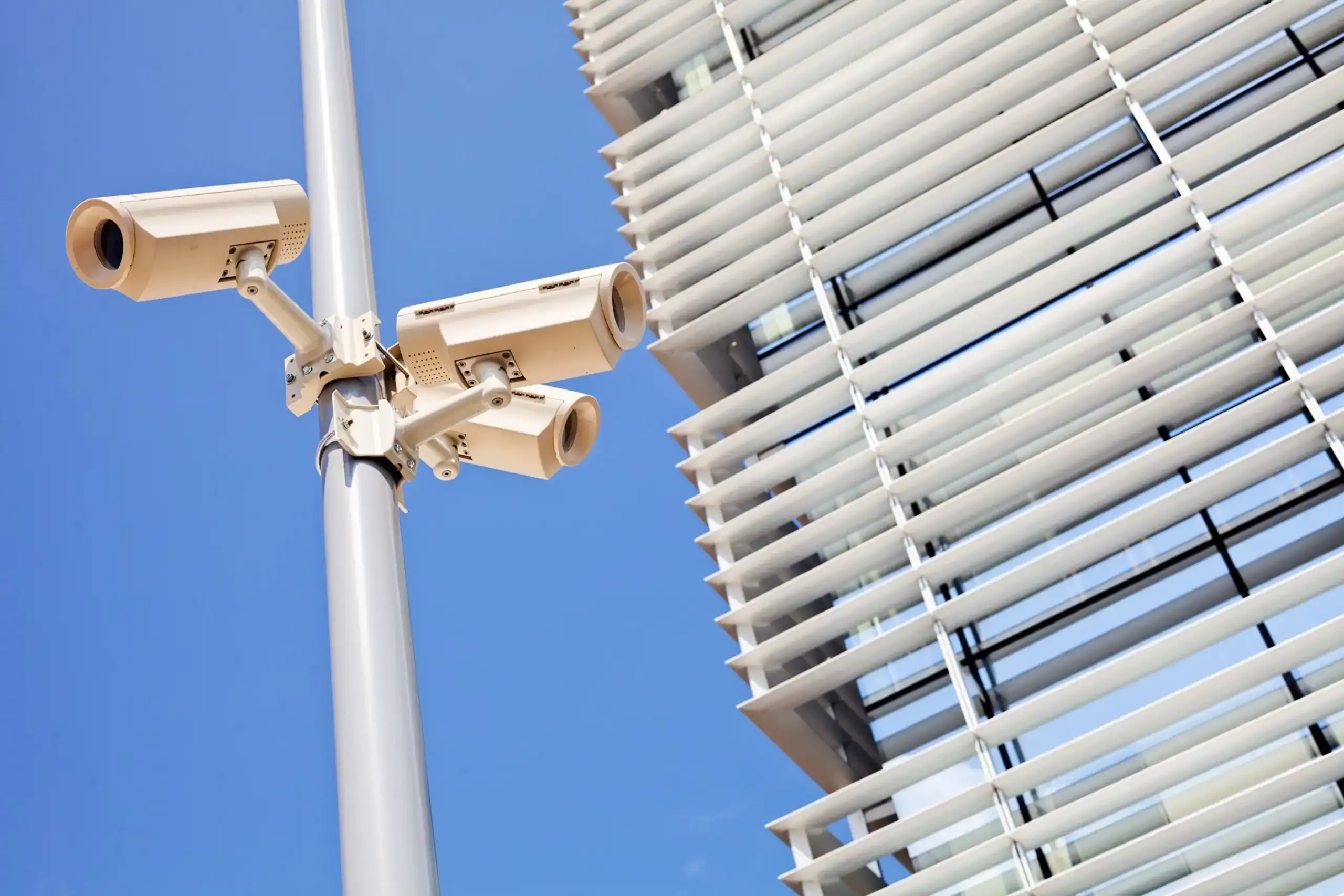 Drei weiße Überwachungskameras an einem grauen Mast vor blauem Himmel, im Hintergrund moderne Gebäudefassade mit horizontalen Lamellen, symbolisiert IT-Security und Netzwerklösungen.