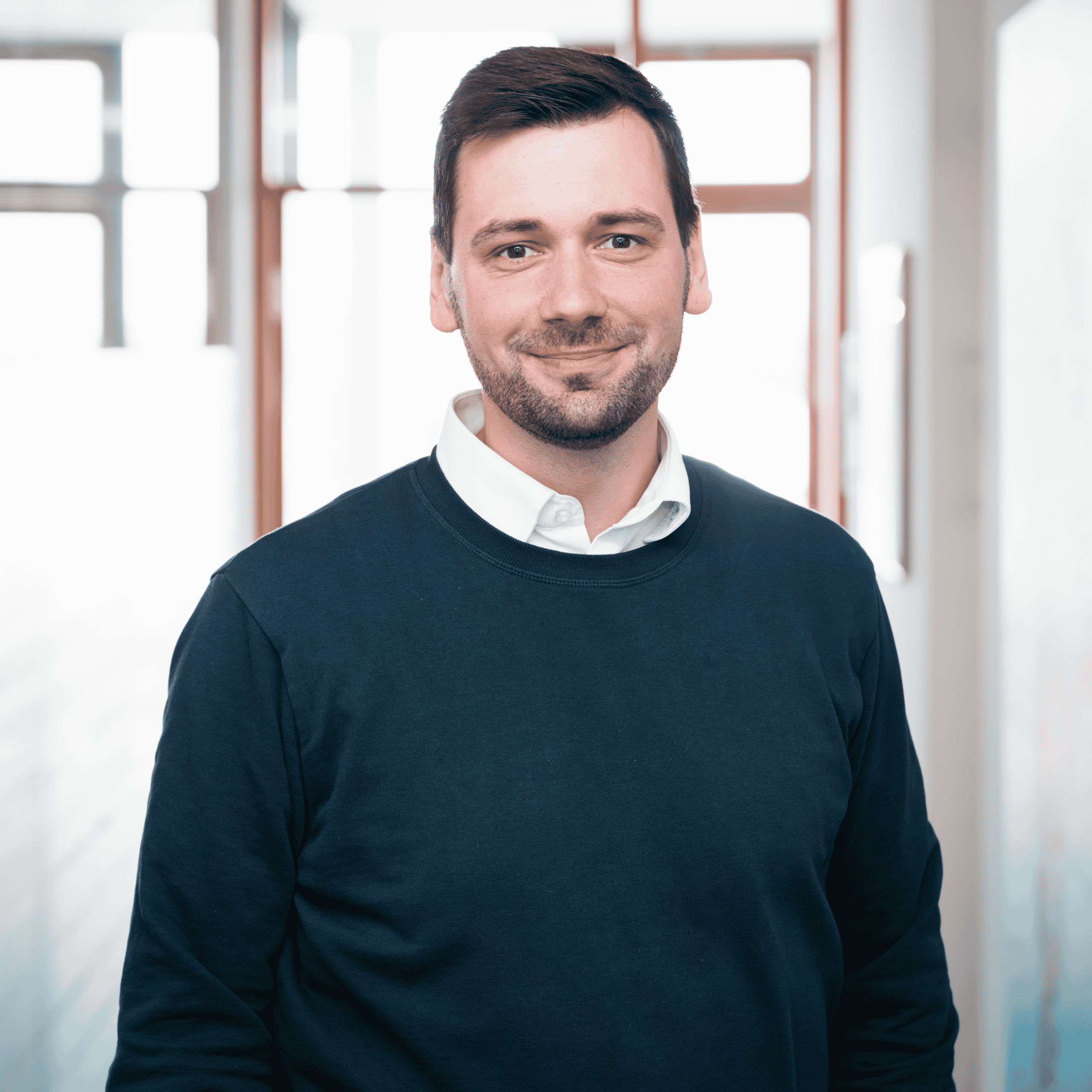 Mann mit kurzem dunklem Haar und Bart trägt weißen Hemdkragen unter dunkelblauem Pullover, steht lächelnd vor hellem Fenster in modernem Büro, symbolisiert freundlichen Ansprechpartner für IT-Beratung und Managed Services bei dierckgroup.