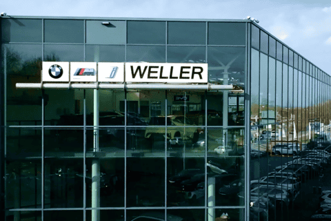 Modernes, mehrstöckiges Büro- und Ausstellungsgebäude mit Glasfassade, auf der Fassade sind die Logos von BMW, M und Weller zu sehen, zahlreiche geparkte Autos entlang des Gebäudes, blauer Himmel mit wenigen Wolken, Umgebung mit Bäumen und weiteren Gebäuden sichtbar.