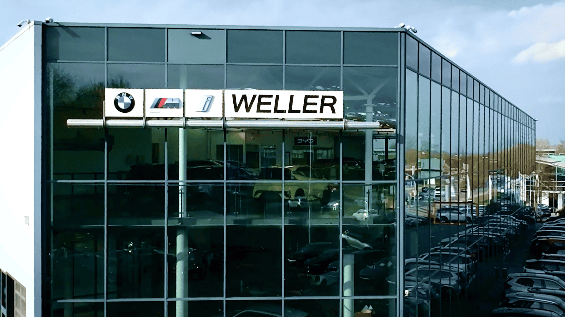 Modernes, mehrstöckiges Büro- und Ausstellungsgebäude mit Glasfassade, auf der Fassade sind die Logos von BMW, M und Weller zu sehen, zahlreiche geparkte Autos entlang des Gebäudes, blauer Himmel mit wenigen Wolken, Umgebung mit Bäumen und weiteren Gebäuden sichtbar.